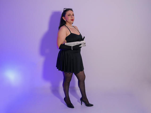 Lisa Taylor está em pé, vestindo um vestido preto, segurando luvas brancas, com meias e saltos pretos. Ela tem cabelo preto em um rabo de cavalo, pele clara e um leve rubor rosa nas bochechas..