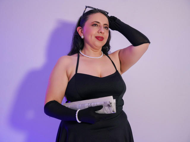 LisaTeylor met een zwarte jurk, parels, zwarte handschoenen, die een krant vasthoudt, haar hand omhoog heeft, een zonnebril op haar hoofd draagt, rode lippenstift, zwart haar, lachend, staand, met een gemiddelde borstmaat, rond, op een witte achtergrond..