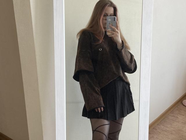 KarKutie with long brown hair, wearing a brown oversized sweater, black pleated skirt, and black patterned tights, taking a mirror selfie with her phone.