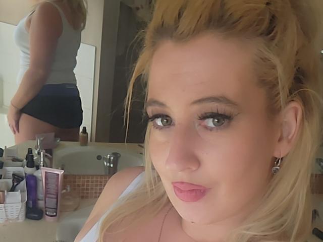 Shh69 has blonde hair, green eyes, and pouty pink lips. She's wearing a white tank top and black shorts, with her ass in the mirror reflection. Bathroom sink visible.
