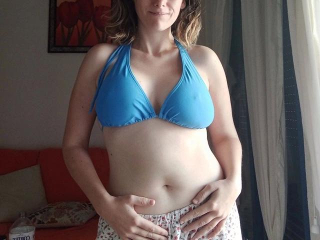 ValerieCalor-hot in a blue bikini top, showing her boobs, with hands on her belly, wearing floral pants, standing in front of a window with white curtains.