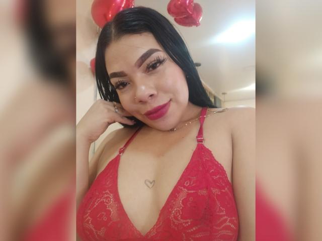 AdrianaPaul con capelli neri, reggiseno di pizzo rosso, tatuaggio a forma di cuore sotto il seno, orecchini a forma di palloncino rossi, rossetto rosso, sorridente, mano sulla guancia, illuminazione soffusa, campo medio, sicura di sé, sexy..