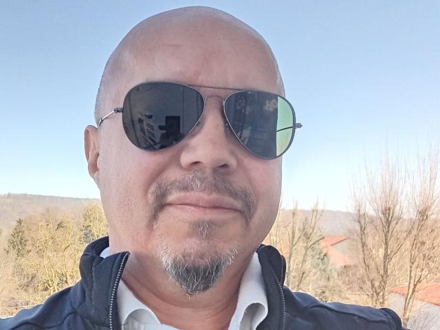 SinfulSmile69 has a bald head, grey goatee, and is wearing black aviator sunglasses with a black jacket over a white shirt. His reflection is visible in the lenses.