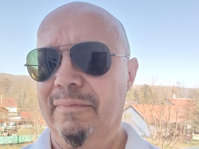 SinfulSmile69 has a bald head, a goatee, and wears dark aviator sunglasses with a white shirt. His face is slightly wrinkled, looking serious.