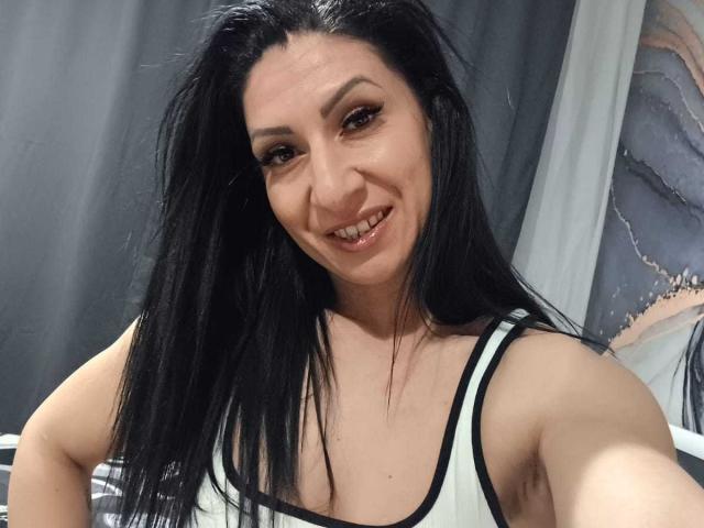 LanceDance heeft zwart haar, bruine ogen en een lachend gezicht met zichtbare tanden. Ze draagt een wit tanktopje met zwarte randen, waardoor haar borsten zichtbaar zijn. Ze maakt een selfie..
