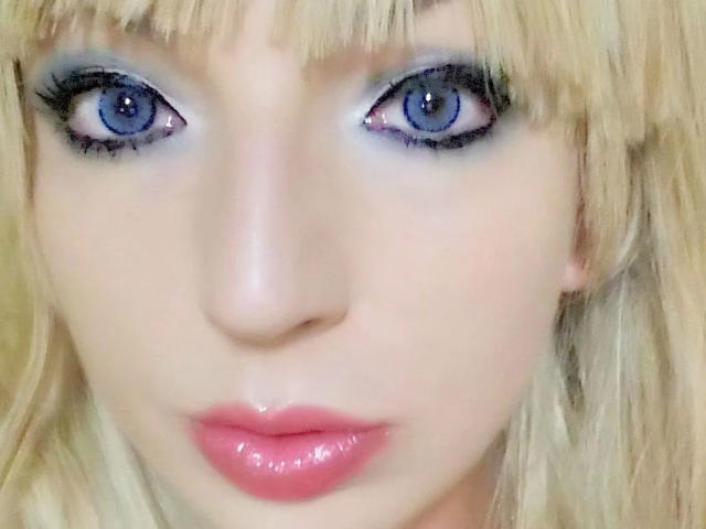 A Lust-Maus-hot tem olhos azuis brilhantes, cabelo loiro com franja e lábios rosados e brilhantes. Ela está olhando diretamente para a câmera com uma expressão sutil e séria..