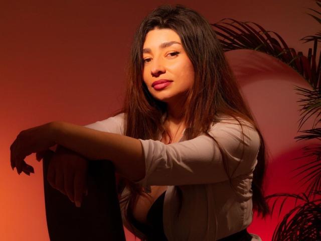 Michonnek with long brown hair, pink lips, and a white shirt, leaning on a chair, arms crossed, looking relaxed and confident, red and orange lighting, subtle smile.