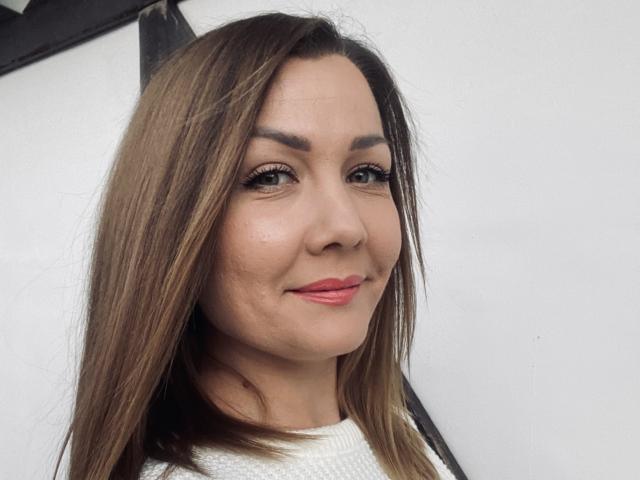 LanaMoon69 has brown hair, light skin, and is smiling with pink lipstick, wearing a white sweater. Her eyes are green with long eyelashes. Boobs are not visible.