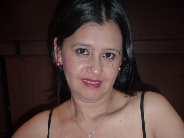 KinkyViolet has medium brown skin, black hair, and pink earrings. She's smiling with red lipstick, wearing a black strap top, and a silver necklace. Boobs are visible.