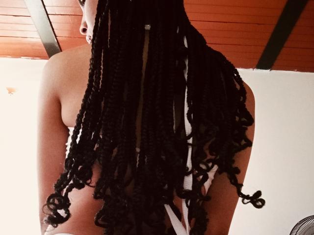 SexyGail's back showing black braided hair, small bare boobs, and a white strap, under a wooden ceiling with green beams. Her skin is dark, and she's topless.