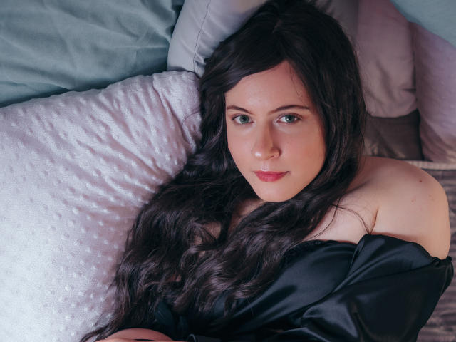 NanaMoonlight has long, wavy dark brown hair, green eyes, and is wearing an off-shoulder black top, lying on a white textured pillow with a subtle smile.