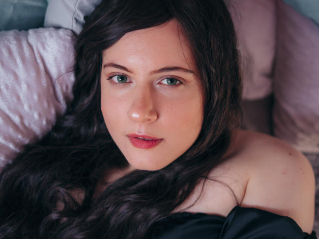 NanaMoonlight has pale skin, green eyes, and dark wavy hair, with slightly parted red lips, lying on pink pillows, wearing a black off-shoulder top, looking directly at the camera.