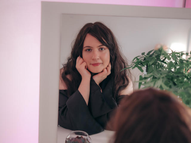 NanaMoonlight with wavy dark hair, black top, hands on cheeks, smiling at mirror, green plant in background, pink light, reflective surface, blurred brown hair in foreground.