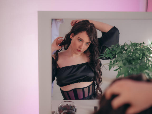 NanaMoonlight with long dark hair, black off-shoulder top, and purple-striped corset, standing in front of mirror, hand in hair, looking at reflection, potted plant on right.