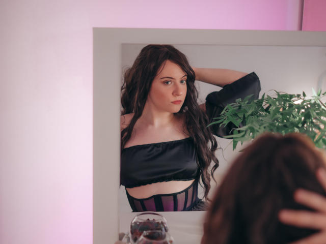 NanaMoonlight, avec ses longs cheveux bruns, se regarde dans le miroir, portant un haut noir à une épaule et des sous-vêtements rayés violet et noir, une main dans ses cheveux. Son reflet montre son expression sérieuse. Une plante en pot se trouve à proximité..