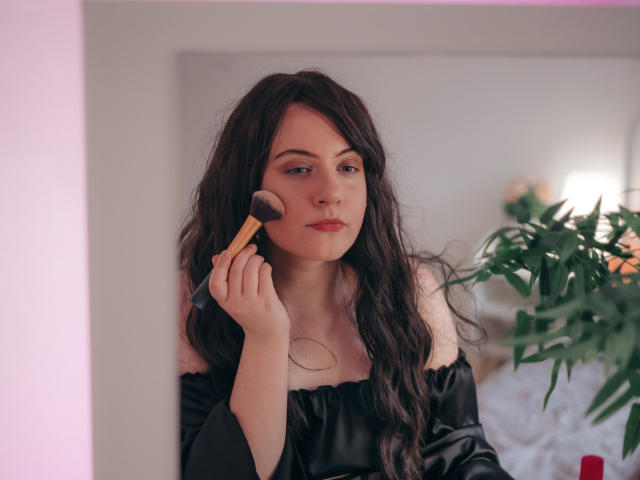 NanaMoonlight, com cabelos escuros e ondulados, blusa preta com um ombro à mostra, aplicando blush com um pincel fofo, iluminação rosa suave, planta verde à direita, expressão calma, lábios levemente entreabertos..
