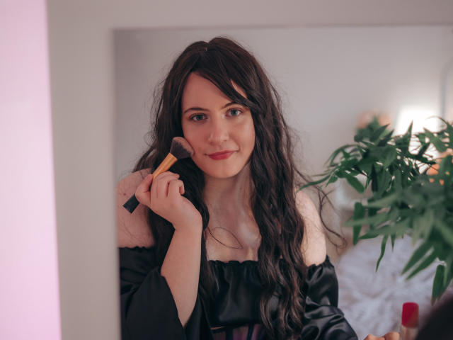 NanaMoonlight with long, wavy black hair, off-shoulder black top, holding a makeup brush to her cheek, smiling gently, standing in front of a mirror.