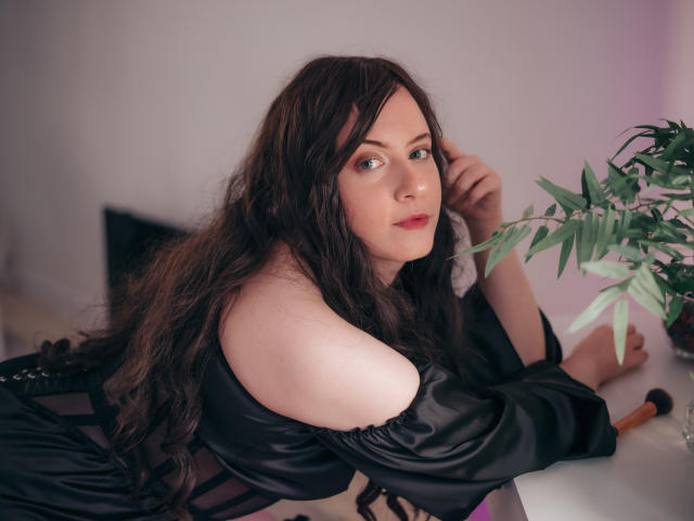 NanaMoonlight with long brown hair, green eyes, and pale skin, wearing a black off-shoulder top, leans on a white table, looking at the camera, with a plant nearby.