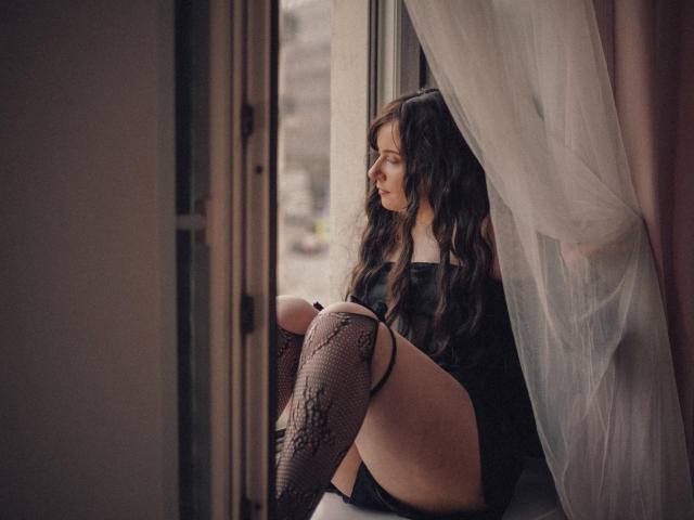 NanaMoonlight, avec ses cheveux bruns ondulés, assise près d'une fenêtre, portant des bas en dentelle noire et un haut noir, regardant à l'extérieur, lumière douce, rideau blanc à droite..