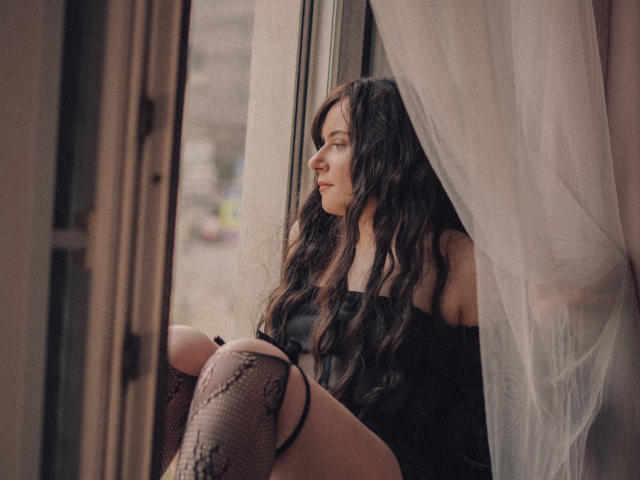 NanaMoonlight, avec ses longs cheveux bruns ondulés, est assise près d'une fenêtre, vêtue d'un haut noir à une épaule et de bas en dentelle, et regarde pensif vers le côté. De rideaux fins et transparents encadrent sa silhouette..