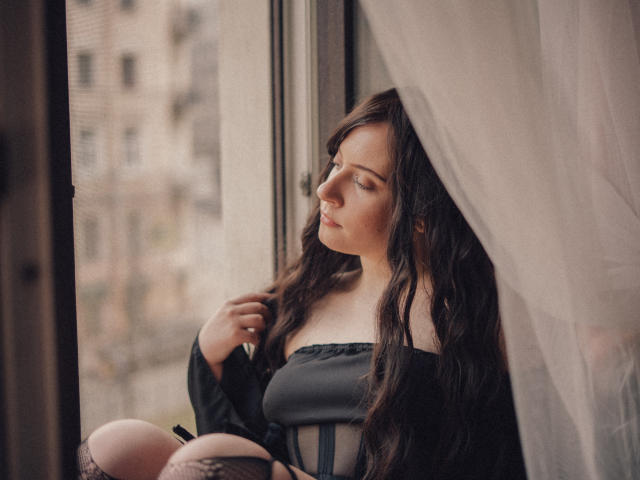 NanaMoonlight, con cabello castaño ondulado, una blusa negra con un hombro descubierto y medias de rejilla, sentada junto a una ventana, mirando hacia afuera, con la mano tocándose el cabello, luz suave, expresión serena..