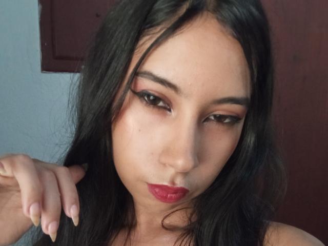 SaraSpecial has dark, wavy hair, red lipstick, and black eyeliner. Her hand is near her face with a serious expression. She has a light brown complexion.