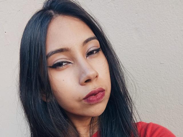SaraSpecial has long black hair, brown eyes with bold eyeliner, and red lips. She's wearing a red shirt, looking slightly to the side with a pouty expression.
