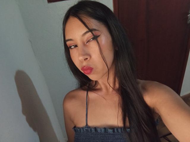 SaraSpecial with long black hair, dark eyes, and red lipstick, pouting her lips, wearing a blue strap top, sitting against a white wall and red door.