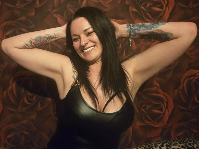 ReifeCleo4U-hot tem cabelo preto, está sorrindo, com os braços levantados, mostrando tatuagens, vestindo uma blusa de couro preta, expondo seios pequenos, com uma pulseira no braço direito..