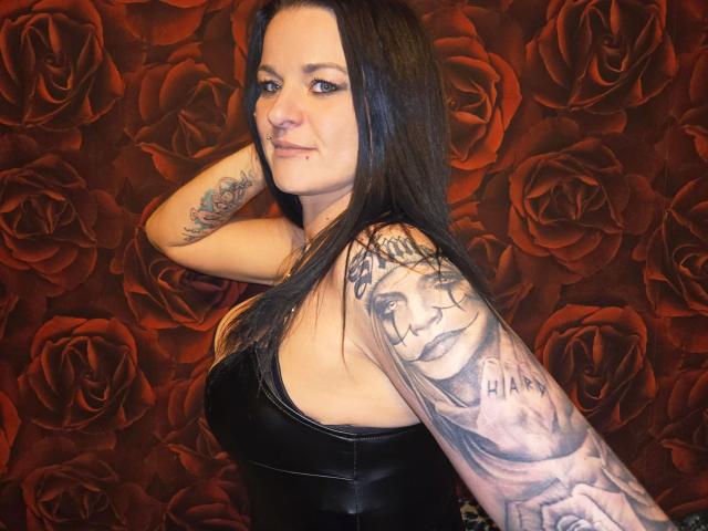 ReifeCleo4U-atractiva, con cabello negro, piercing en la nariz y tatuajes, vistiendo una blusa de cuero negro, sonriendo, mostrando un tatuaje detallado en el brazo sobre un fondo con estampado de rosas rojas..