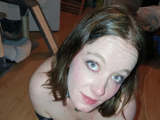 DollySoumise69 has pale skin, blue eyes, brown shoulder-length hair, and is looking up with a slight smile, showing her small tits and a black strap above her chest.