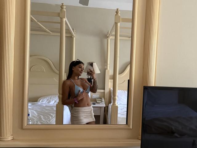 ThaliaAres en un selfie frente al espejo, vistiendo un sostén azul y una falda blanca, mostrando sus senos y un tatuaje en su brazo, de pie en un dormitorio con una cama con dosel..