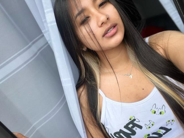 Ammy Lewis, con lunghi capelli neri e biondi, indossa una canotta bianca, ha un piercing al naso ed è rivolta verso la fotocamera con un leggero sorriso, mostrando i suoi piccoli seni..
