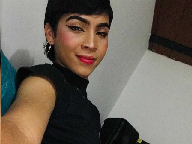 SexyGail has short black hair, thick eyebrows, red lipstick, and is wearing a black sleeveless top with a high neck, holding a black bag, looking at the camera.