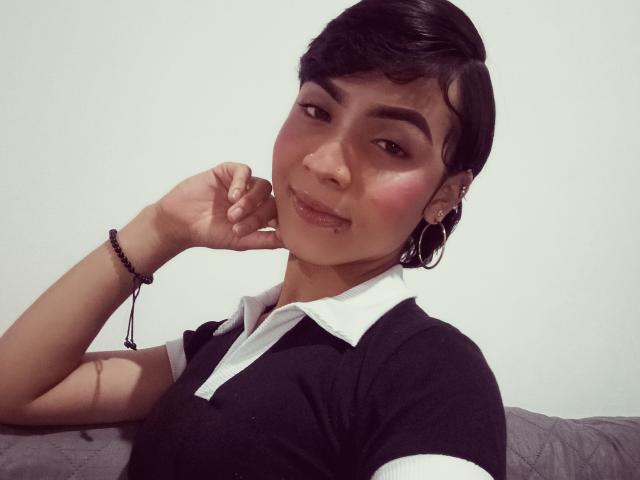 SexyGail with short black hair, blushing cheeks, and a cute smile, wearing a black and white polo shirt, resting her chin on her hand. She's got a bracelet and earrings.