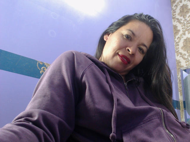 LizethBells, con cabello negro largo, vistiendo una sudadera morada, lápiz labial rojo, sonriendo ligeramente, tomando un selfie desde un ángulo bajo, con sus senos parcialmente visibles, en una habitación con paredes moradas..