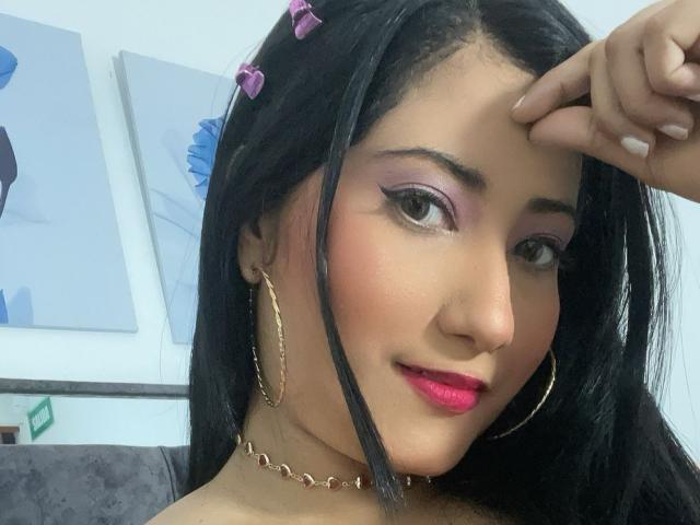 AlexaCooper with pink eyeshadow, pink lipstick, black hair, gold hoop earrings, chain necklace, purple hairpins, and a slight smile, touching her forehead.