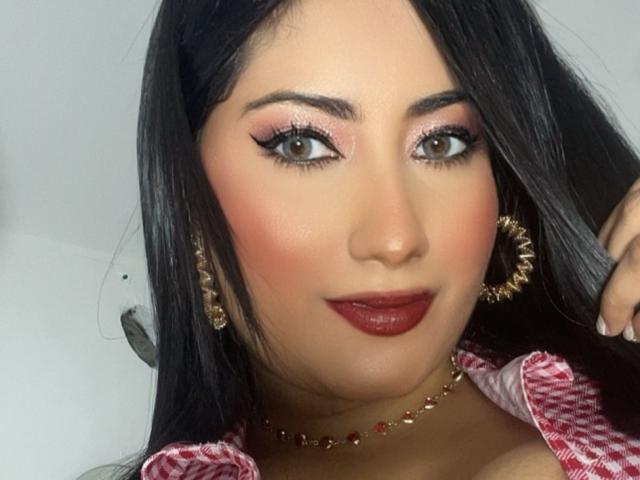 AlexaCooper has tan skin, long black hair, pink eyeshadow, thick black eyeliner, red lipstick, gold earrings, and a red checkered shirt, showing her tits slightly.