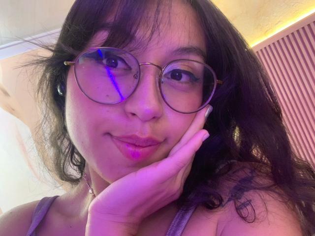 MadyJackson with puffy pink lips, wearing round glasses, black wavy hair, touching her cheek, wearing a grey tank top, showing a cute smile, small tattoo on her right shoulder.