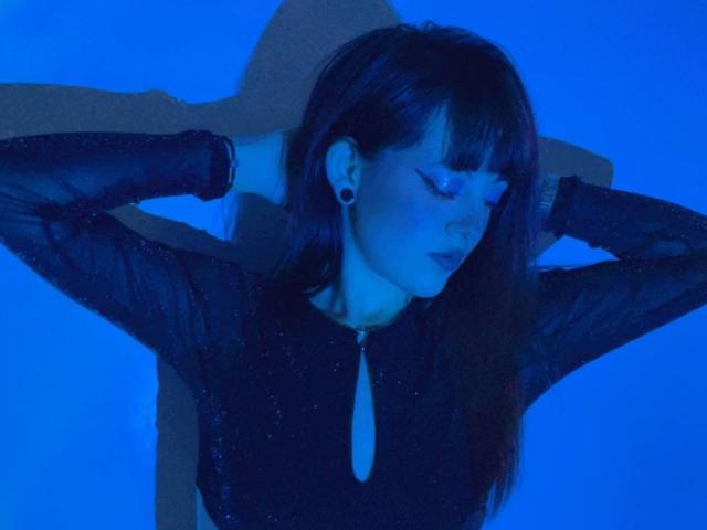 IsabellaKissy, con las manos en el cabello oscuro, vistiendo una blusa negra transparente con cremallera, iluminada en azul, mirando hacia abajo, con aretes grandes, y la sombra de una cruz detrás de ella..