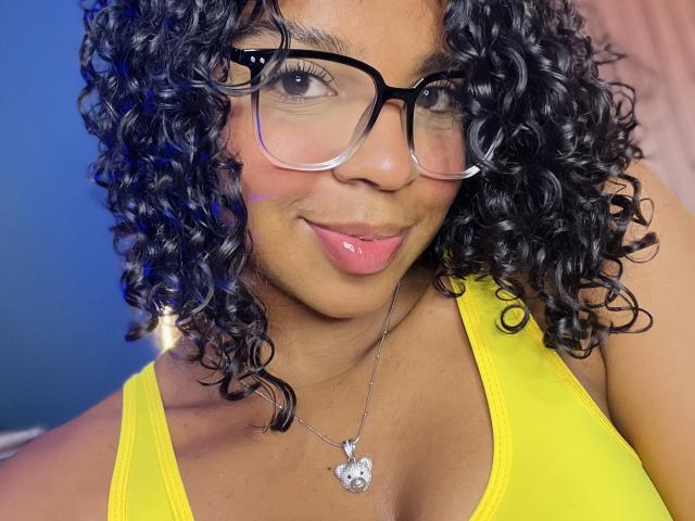 LisaMendez with curly black hair, wearing glasses, yellow tank top, and silver necklace with heart-shaped pendant, showing pink lips and slightly smiling. Boobs visible in yellow top.