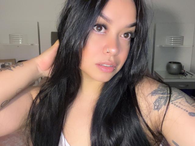 Laureenn with long black hair, brown eyes, nose ring, and tattoos on her arms, looking at the camera, wearing a white top, showing her small tits, and shiny lips.