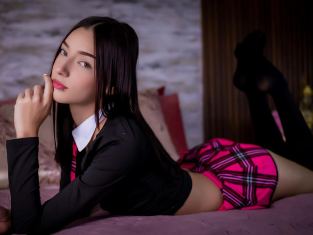 AliceBakerr, with long black hair, lies on a bed in a pink plaid skirt and black top, her finger touching her pink lips, wearing black socks.