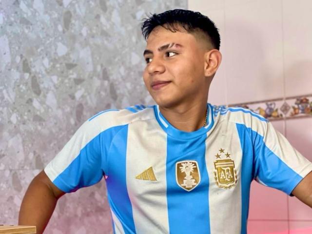 RichiMontes, con cabello negro corto, vistiendo una camiseta de fútbol argentina azul y blanca, sonriendo ligeramente, mirando hacia un lado, de pie en una habitación con una pared gris texturizada..
