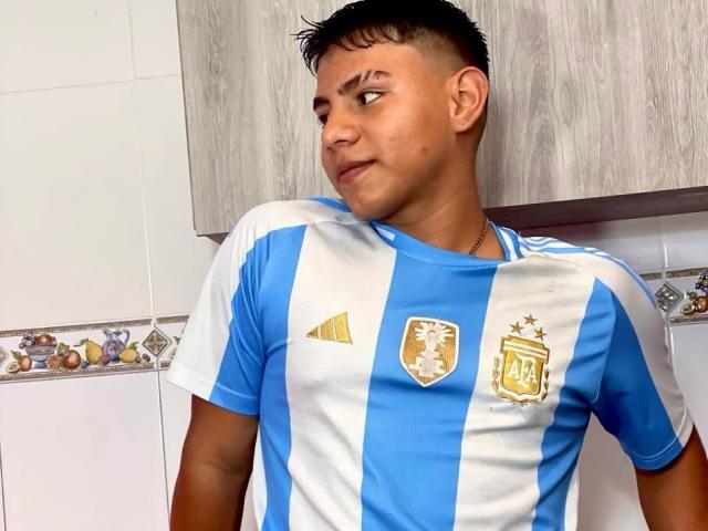 RichiMontes, kort zwart haar, bruine huid, draagt een blauw en wit Argentijns voetbalshirt met een gouden embleem en een diamantpatroon, staat, kijkt zijwaarts, met een lichte grijns, een houten kast op de achtergrond..
