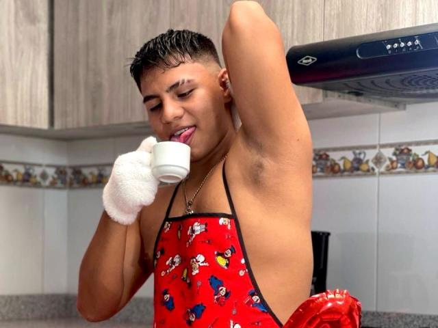 RichiMontes, con el cabello negro y húmedo, delantal rojo, lengua afuera, lamiendo una taza blanca, con la axila visible, usando un guante blanco, en la cocina, sobre la estufa, sonriendo..