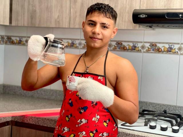 RichiMontes, musculoso, com pele bronzeada, cabelo curto e escuro, vestindo um avental vermelho com estampas de desenhos animados, luvas brancas, segurando um pote de vidro e uma caneca branca, sorrindo em uma cozinha..