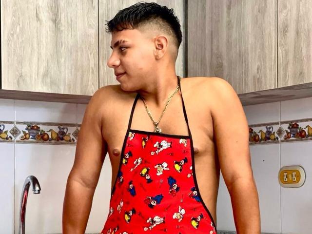 RichiMontes con un delantal rojo que presenta personajes de dibujos animados, sin llevar nada más, mostrando su pecho desnudo, musculoso y de piel bronceada, y su cabello corto y oscuro, de pie en una cocina..