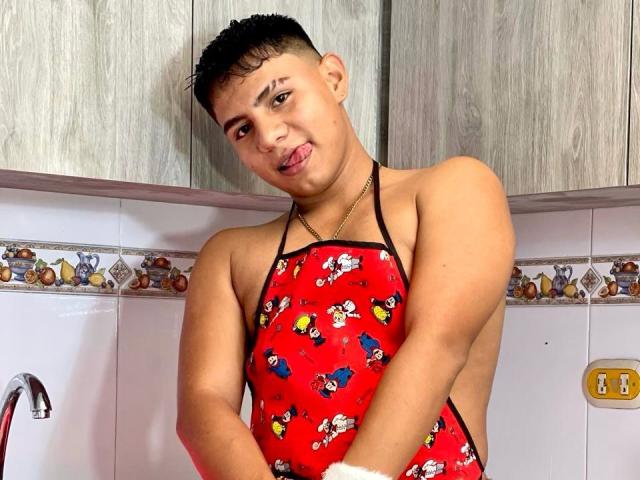 RichiMontes, sin camisa, vistiendo un delantal rojo con estampados de Mickey Mouse, sacando la lengua, cabello negro corto, piel clara, de pie en una cocina, con las manos cerca de sus glúteos..