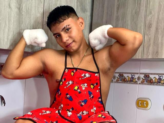 RichiMontes, sin camisa, vistiendo un delantal rojo con personajes de dibujos animados, mostrando sus bíceps, con guantes blancos, cabello negro corto, piel marrón claro, pezones pequeños, sentado en una cocina..